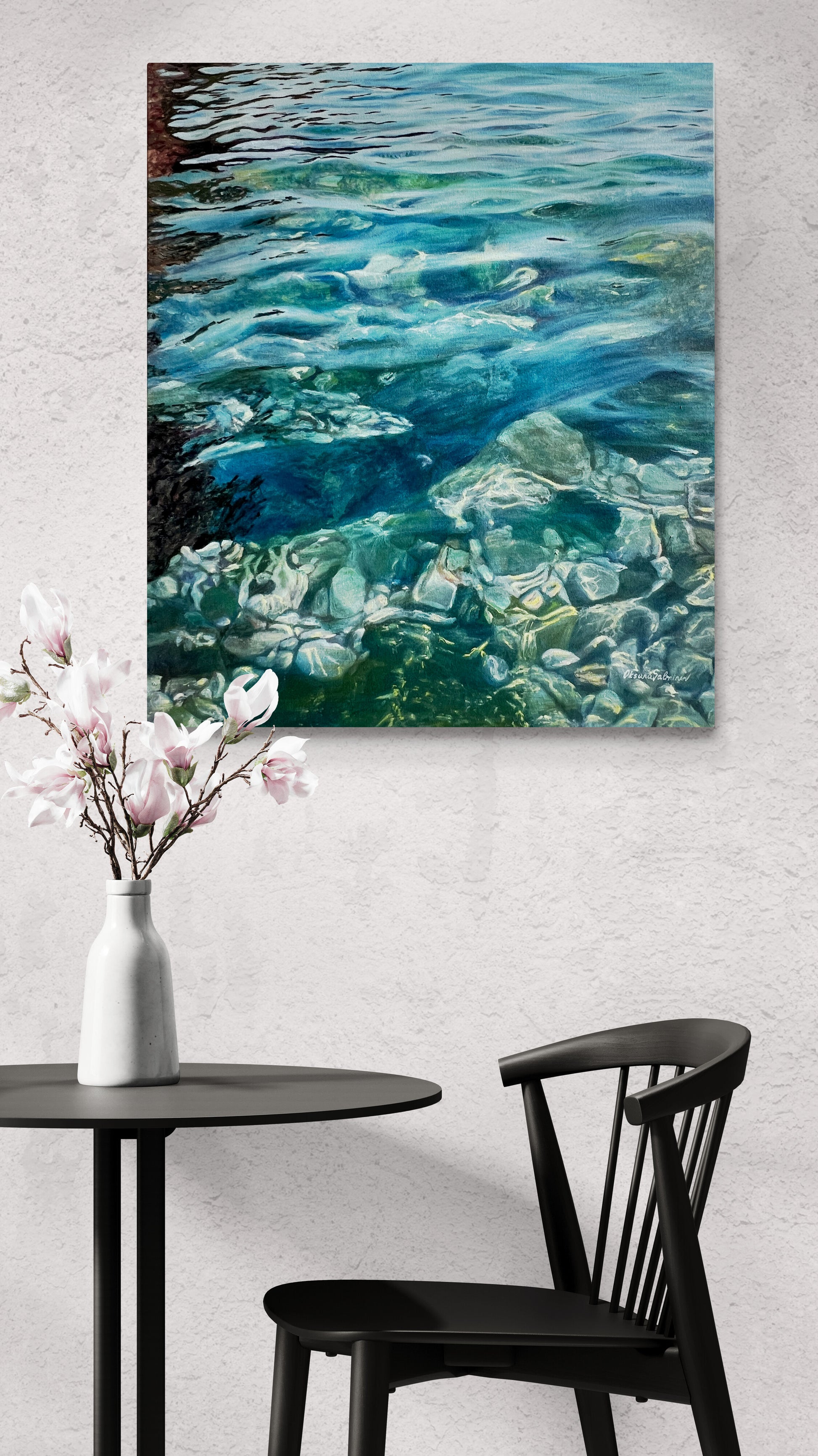 The painting of water and stones on a wall above a table with a vase and chair.