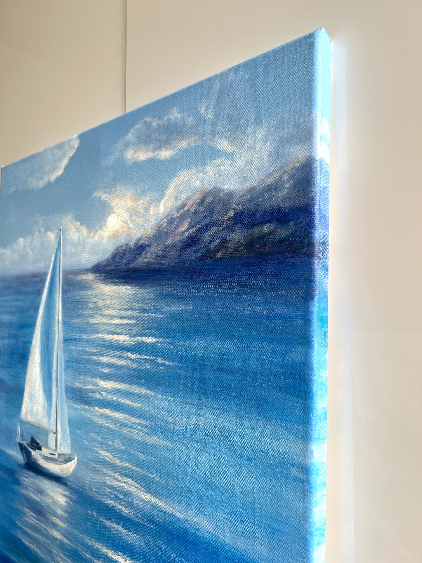 A canvas painting of a sailboat on a blue sea with mountains and clouds, shown hanging against a light wall.