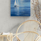 A square canvas painting of a white sailboat on a blue sea with mountains in the distance, displayed on a textured light gray wall above a modern wicker chair.