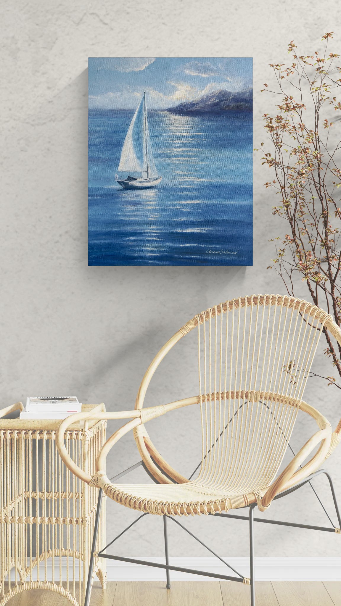A square canvas painting of a white sailboat on a blue sea with mountains in the distance, displayed on a textured light gray wall above a modern wicker chair.