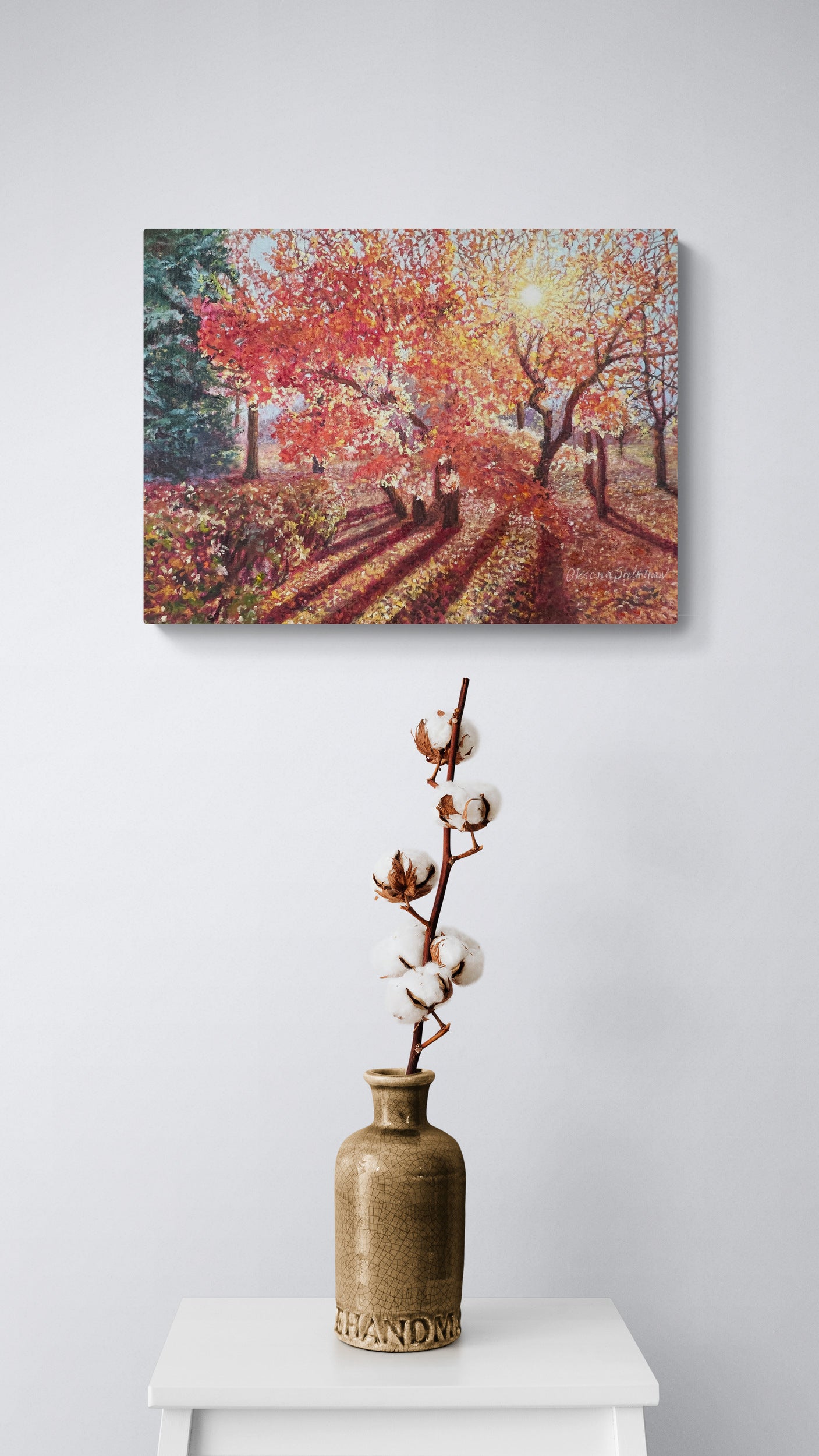 Oil painting of a colorful autumn landscape above a vase with cotton branches on a white surface.