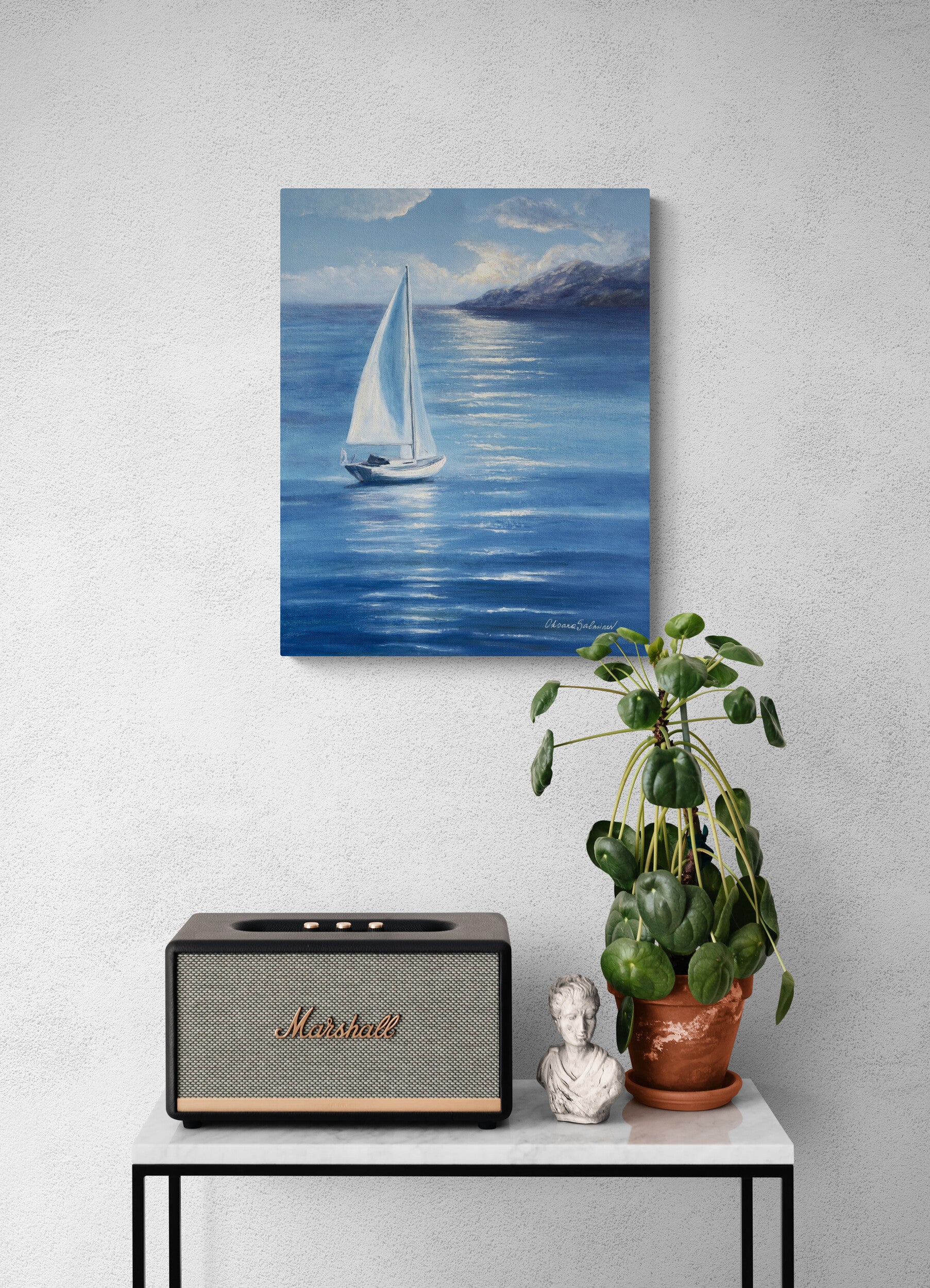 A vertical canvas print of a blue seascape with a white sailboat, displayed on a textured light gray wall above a console table with a Marshall speaker and a potted plant.