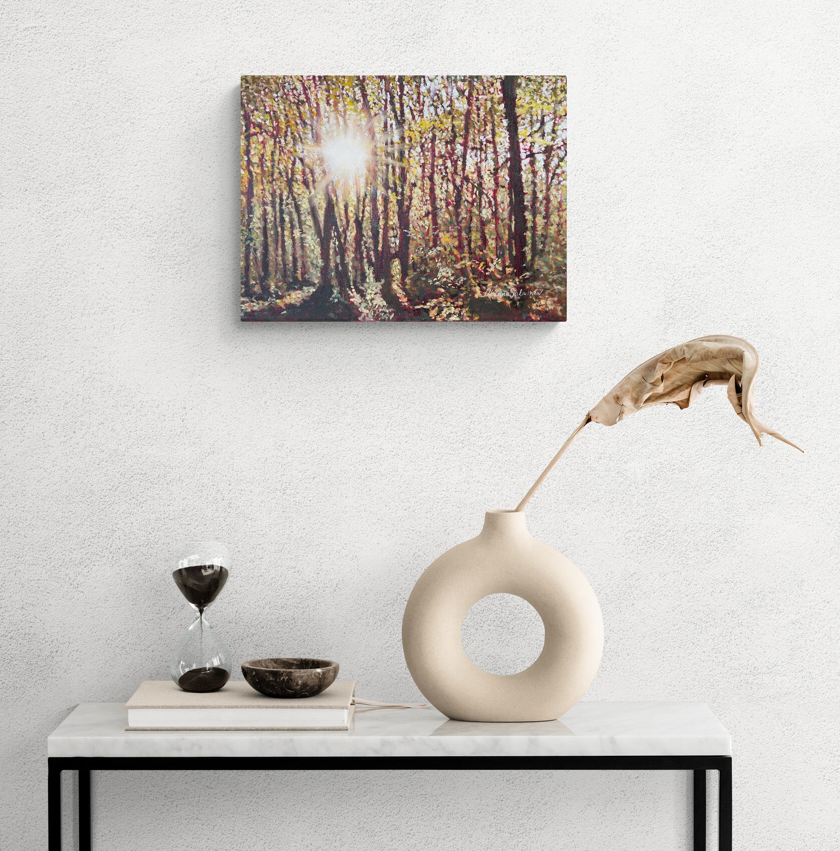 Decorative setup with a vase, hourglass, and bowl on a table against a wall with a forest painting.