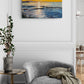 Original oil painting on canvas of a sunset over the ocean hangs on a white paneled wall above a gray armchair in a styled living room.
