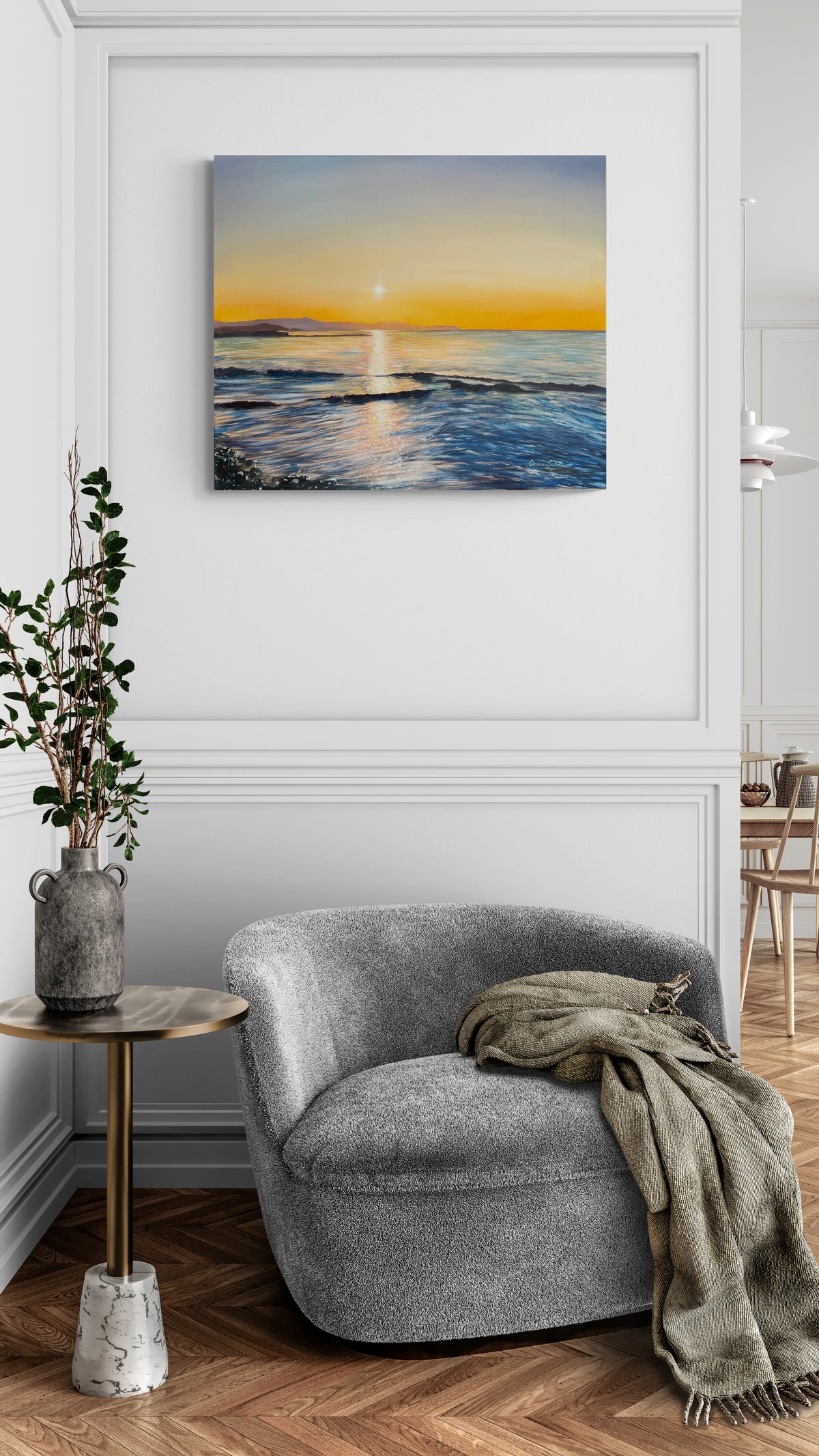 Original oil painting on canvas of a sunset over the ocean hangs on a white paneled wall above a gray armchair in a styled living room.