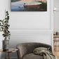 Unframed painting of a boat on a lake above a gray armchair with a blanket in a room.