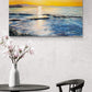 A square canvas painting of a sunset over the ocean hangs on a textured white wall above a black table with a white vase of pink flowers and a black chair.