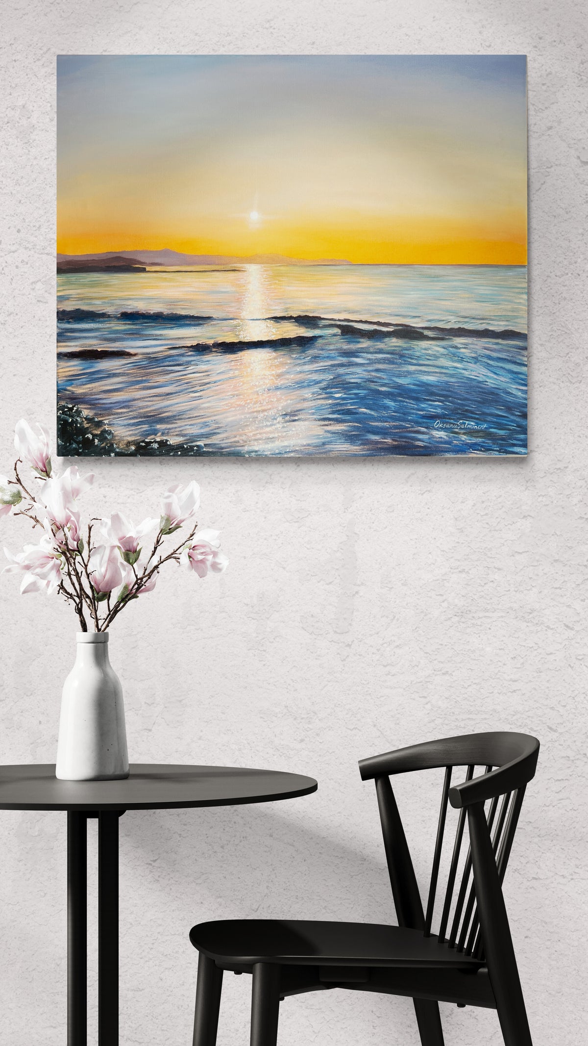 A square canvas painting of a sunset over the ocean hangs on a textured white wall above a black table with a white vase of pink flowers and a black chair.
